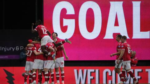 Wrexham players celebrate in a huddle in front of a large scoreboard with the words 'Goal'