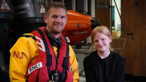 Andy Read, wearing a yellow RNLI jacket and red life jacket, and Billie, who has a black top, smile at the camera.