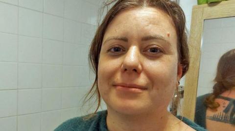 Headshot of Sam in her bathroom. She has wavy brown hair tied back, with some strands framing her face. She is wearing a green off-the shoulder jumper - and reflected in the mirror behind her you can see it is tied at the back and she has a tattoo. The wall behind her has white tiles, there's a shower curtain and wooden framed bathroom cabinet.