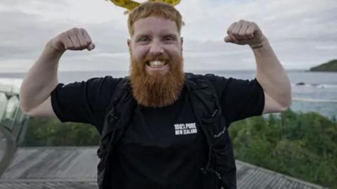 Russ Cook flexing his arm muscles as he stands in front of a yellow sign and a body of water. He is wearing a black t-shirt and looking directly at the camera.