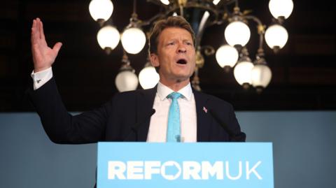 Reform UK Deputy Leader Richard Tice in a suit and blue tie addressing a press conference in London. In front of him is a blue lectern that reads: Reform UK