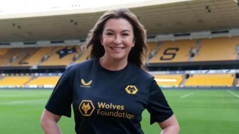 Suzy is on the pitch at Wolves wearing a black Wolves Foundation t-shirt. She is smiling and has long dark hair.