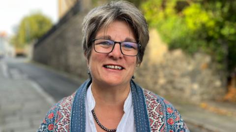 Deputy Jayne Ozanne is standing by a road. She has grey hair and a pair of black rectangular rimmed spectacles, while wearing a patterned coat on top of a white shirt.
