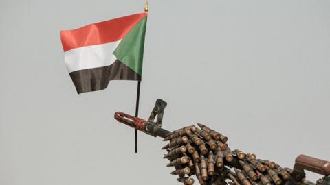 A Sudanese flag placed above the muzzle of a machine gun covered with an ammunition belt.