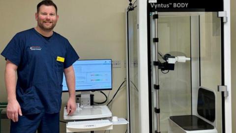 A man wearing navy blue medical scrubs. He is standing next to diagnostic equipment and is smiling for the photograph.