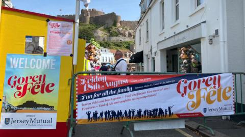 A picture of the entrance to the fete. There is a metal barrier on which are posters promoting the event. There is also a shop in the background with people milling around.
