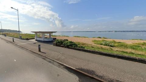 Road and path with beach and water beyond and an art deco shelter standing in the centre.
