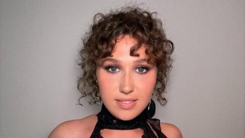A girl with brown curly hair, put up into a classy messy bun, she's in a black halter neck dress, dressed for a special occasion. She is wearing makeup with smokey eye shadow and pink lips. Her eyes are a dark blue which makes the eyeshadow pop. 
