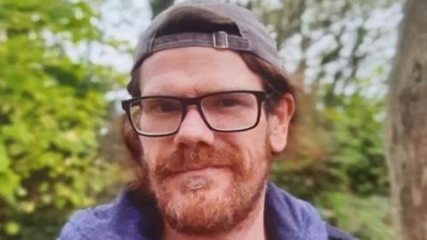 Shane Lynch is smiling at the camera as he stands outside with foliage behind him. He has a ginger brown beard and collar-length hair. He is wearing a baseball cap backwards and a pair of brown rimmed glasses.
