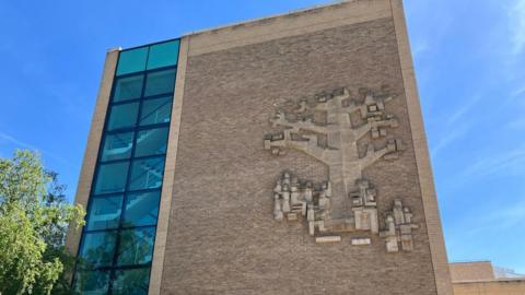 View from the road looking up at the building on a sunny day. It is tall with light bricks and glass from the ground floor up, on the left side of the building, to show a staircase. On the right side is the large sandstone sculpture about the height of four of the short staircases . It is blocky and shows people sitting under an oak tree. 