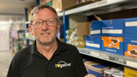 A man in glasses, with short hair and stubble is wearing a black polo shirt with a changing lives together on it. There are racks of food behind him