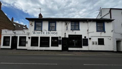 A large, white tow-storey pub beside a two-lane road. Ther are cloudy skies above it. The pub has 12 large windows and two doors. The pub's name, "The White Horse" is written on the front.