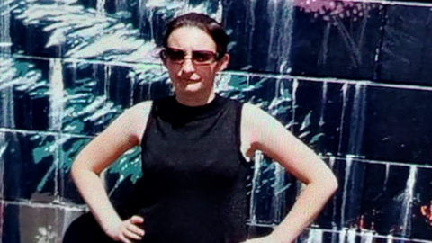 Woman wearing sunglasses wearing a black tunic with her hands on her hips, standing in front of a wall