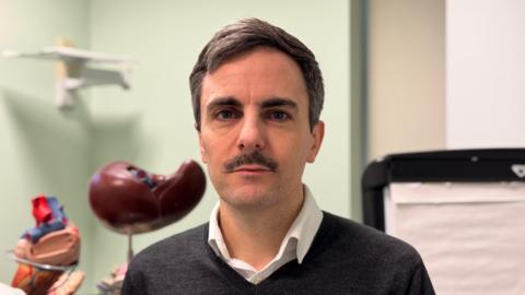 Tom Dolphin, a man with brown hair and a moustache, wears a grey jumper and a white shirt.