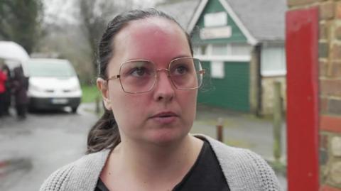 A woman wearing glasses, a black top and a grey cardigan. There is a white van in the background. 