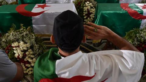 An Algerian man salutes in front of coffins draped in the national flag