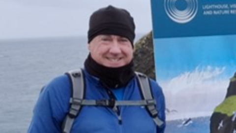 A smiling Ian Napier wearing an outdoor jacket and hat with a rucksack over his shoulders