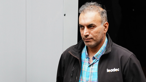 A man in a blue checked shirt and black jacket walking out of a court with a white and black background