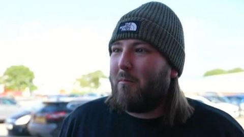 A man with a beard is wearing a hat and a dark top. A car park is behind him.