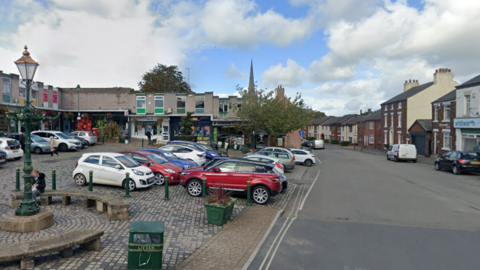 Market Square in Kirkham. The parking area is cobbled with room for about 20 cars. There are rows of shops to the left of the square, and a road and some houses and more shops to the right. There are curved benches in front of the car park for shoppers to sit on.