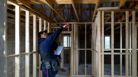 Wide shot of two male builders working on a property. They are wearing work clothing, working together to manage the project. The frame of the new build is half complete around them.