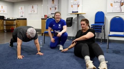 Two women are doing exercised on a carpeted floor. A woman wearing a football jersey crouches between them. 