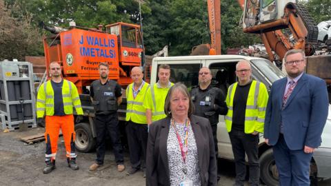A group of council workers in high vis jackets, two police officers and a man and a woman in suits are stood by a heap of crumpled metal in a scrap yard