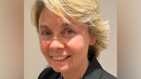 Sam Ridding, a woman with blonde hair and wearing a black shirt is smiling. She is standing against a light-coloured background.