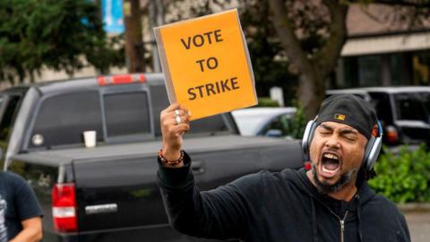 A man wearing a baseball cap backwards and headphones on shouts and holds up a placard saying 'vote to strike'
