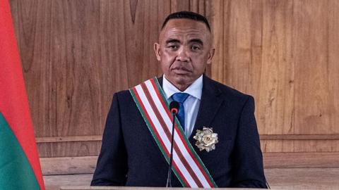 Randrianirina standing wearing a black suit and a green, red and white striped sash, with an official badge on his left lapel. there is a mic in front of him and a partial Malagay flag to his right.