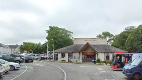 Google image of Holsworthy Memorial Hall. The building is cream. Cars and buses are parked in the car park outside of the hall. Green trees are behind the building. 