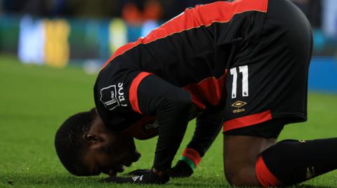 Dango Ouattara wearing red and black Bournemouth kit kneels on a football pitch, forehead touching the grass
