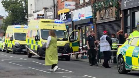 Image shows a street closed off, with paramedics and their vehicles standing on the pavement with members of the public. Red and white incident barrier tape can be seen around one of the ambulances. 