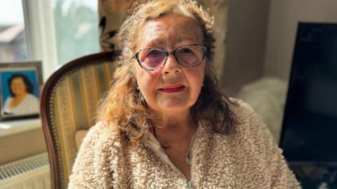 Millie Blenkinsop-French  has shoulder length wavy brown hair, worn half up. She has red lipstick on and wears reading glasses. She's sat on a chair in her living room and is wearing a cream fleece.