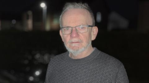 Ciarán O'Doherty is wearing an aron style jumper. He is standing at an EV charging point, it is night time and very dark