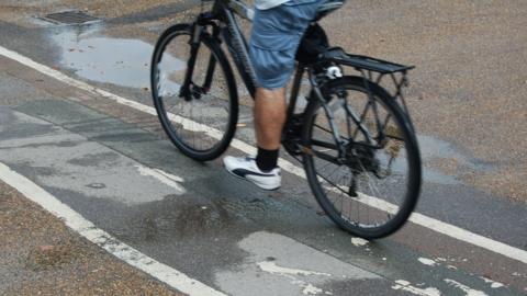 A man cycling on a cycle path. Only the lower half of his body is visible. He is wearing blue shorts, black socks and white trainers.