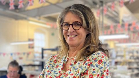 Emma Bridgewater - a woman wearing a patterned dress, glasses and a pearl necklace - is standing with her arms folded. She is smiling for the camera. Behind her is a factory with union jack bunting.
