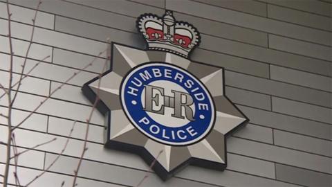 The Humberside Police badge on the wall of a police station. It has the words "Humberside Police" in white letters in a circle with a blue background and the initials "ER" in the middle. A crown sits on top of the badge.