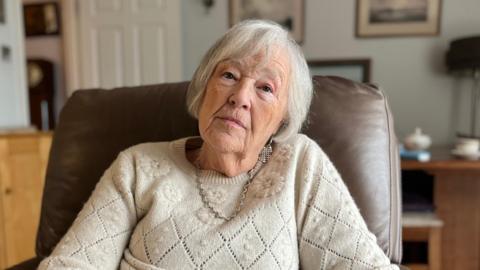 A grey-haired woman sits in a leather arm chair and looks solemn into the camera. She has a beige jumper on. 