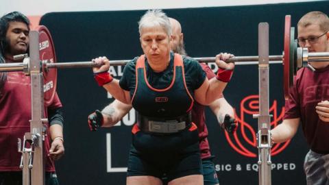 Dee Atkins-Greig in a powerlifting competition. The barbell is on her shoulders, her face shows effort.