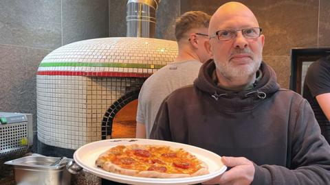 Mark Clarey smiling as he holds a handmade pizza. Behind him is a pizza oven