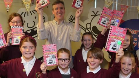 Kinney is at the back smiling and holding up copies of his books. He is with seven smiling children in maroon jumpers also holding up his books. In the background are cartoons of the wimpy kid from his stories. 