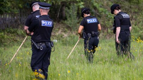 Four police officers, two men and two women, in black uniforms stand with their backs to the camera in a patch of long grass carrying wooden sticks used to search for weapons or drugs. 