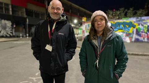 A man and woman dressed in thick coats stand on a dark street