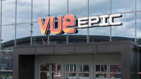 A close-up of a sign fixed to a glass building, which reads VUE EPIC. Below the sign are the modern glass doors into the cinema building.