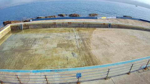 An empty concrete pool with a 'NO DIVING' warning painted near the bottom edge. A metal railing surrounds the pool, and a ladder is visible on the far side. In the background, the ocean stretches out with several rocks protruding from the water.
