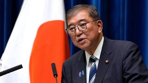 Shigeru Ishiba wearing a dark suit, white collared shirt and striped blue and white tie speaks at a podium in front of a blue curtain and a large Japan flag