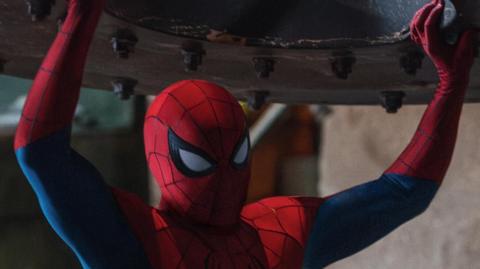 An image from Spider-man Brand New Day where the titular hero, dressed in red and blue skintight suit with big white eyes and black outlines, holds a massive metal disc over his head.