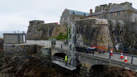 Engineers wearing safety helmets and high-vis jackets are stood on scaffolding looking at a bridge leading up Castle Cornet in Guernsey.