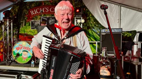 Accordian player from the Wurzels wearing a red scarf on his neck singing at the camera with the drummer in the background